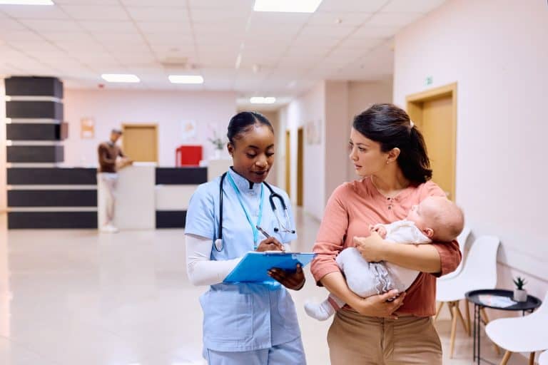 Black nurse writing data in medical record while talking to mother with baby at doctor's office.